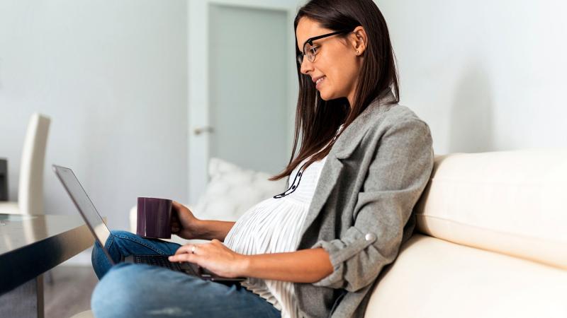 A pregnant women holding a mug and looking at a laptop computer while sitting on a couch. 